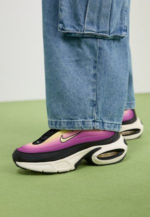 Fioletowo-żółto-czarne sneakersy Nike noszone z niebieskimi jeansami na jasnooliwkowej podłodze.