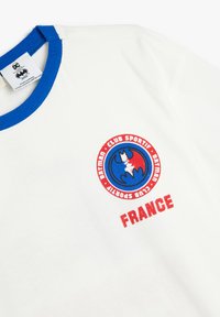 Camiseta blanca con cuello de canalé azul. Presenta un gráfico redondo con un murciélago estilizado y "FRANCIA" en rojo, añadiendo contraste de color. Material de algodón.