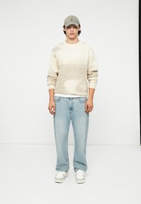 Muška kremasta džemper s uzorkom, svijetloplave traperice i bijele tenisice. Džemper ima mekanu teksturu i opušteni kroj, s rebrastim manžetama.