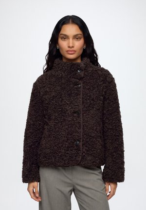 Mujer con cabello largo y oscuro lleva una chaqueta marrón oscuro con textura y botones, y pantalones grises, de pie frente a un fondo blanco liso.