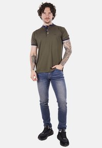 Polo verde oliva con colletto blu navy e righe bianche, abbinato a jeans blu e sneaker nere. Presenta maniche corte e patta con bottoni.