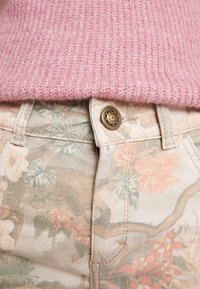 Pantalones de algodón estampados en beige con flores, con acentos en verde y melocotón, que presentan un botón dorado y detalles de costura, combinados con un suéter de punto rosa.