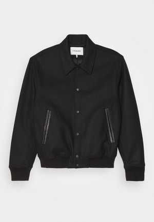 Mężczyzna w czarnej zapinanej na guziki kurtce, ciemnym swetrze, niebieskich dżinsach i białych tenisówkach, stojący z jedną ręką w kieszeni na jednolitym tle.