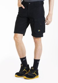 Shorts cargo noirs avec poches latérales, en tissu résistant, arborant un petit écusson jaune. Associés à des baskets noires et des chaussettes.