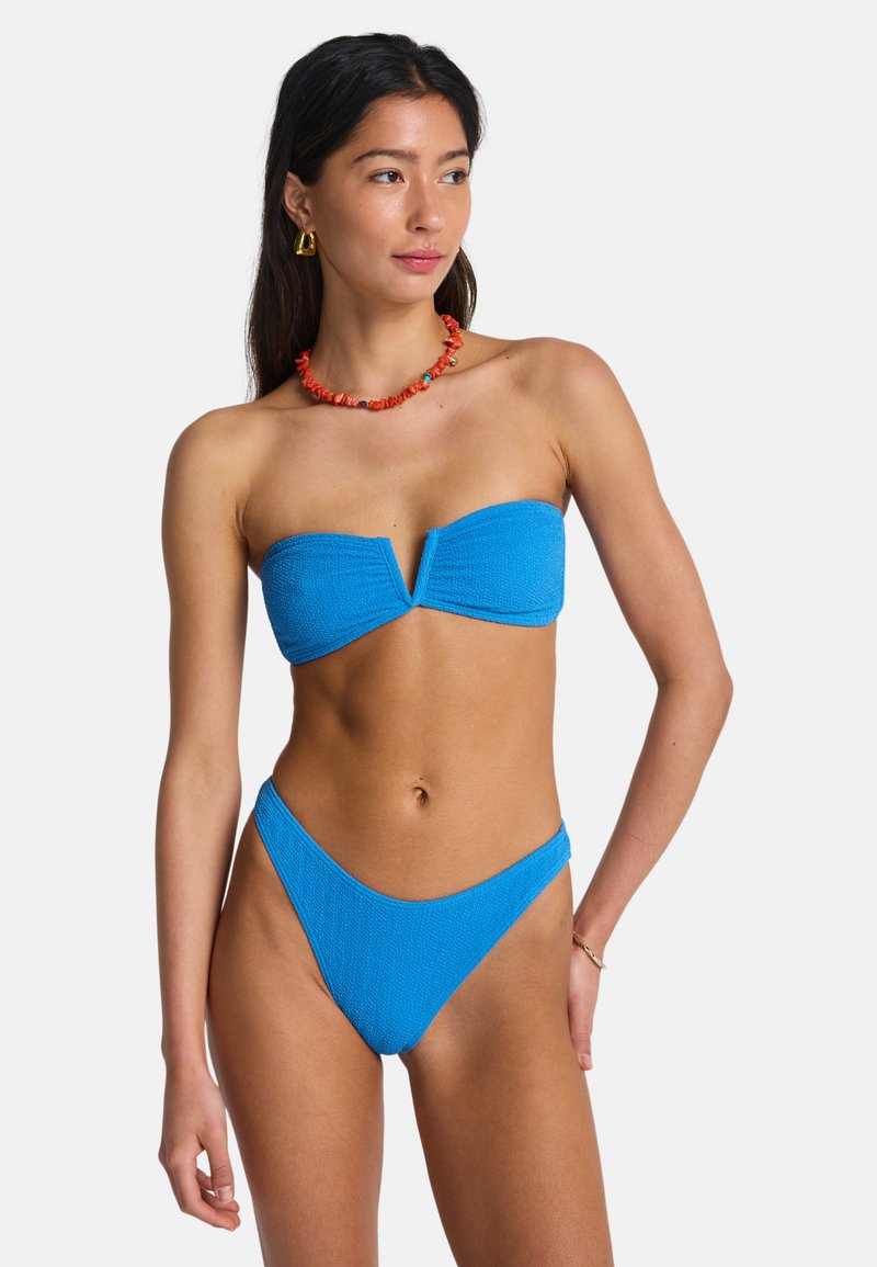 Niebieski teksturowany bikini bandeau z dekoltem w kształcie litery V i pasujące majtki z wysokim stanem. Noszone z koralowym naszyjnikiem z koralików i złotymi kolczykami.
