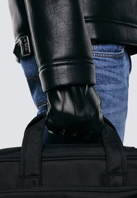 Czarna sk órzana rękawiczka trzymająca czarną materiałową torbę. Osoba ubrana jest w fakturowaną czarną skórzaną kurtkę i niebieskie dżinsy, o prostym, wytrzymałym fasonie.