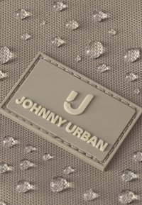 Vanddråber på tekstureret beige stof med en syet beige label med teksten Johnny Urban og en stiliseret J-logo.