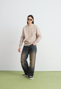 Felpa beige con colletto rotondo, leggera usura sui jeans blu e sneakers scure. Il modello indossa occhiali da sole rettangolari. Sfondo di pavimento verde.