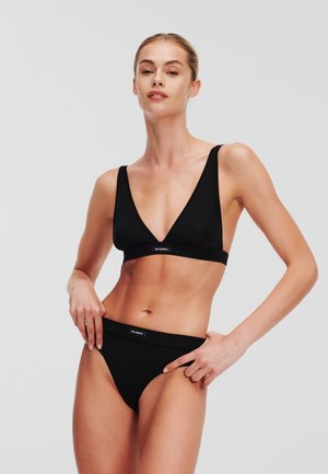 Vrouw in zwart bikiniset met driehoekige top en hoog uitgesneden broekje, poserend voor een effen lichtgrijze achtergrond.