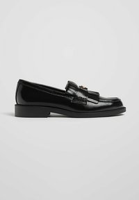 Mocassin en cuir noir avec un petit talon, détails de frange et de gland sur le dessus, présenté en profil latéral sur un fond clair.