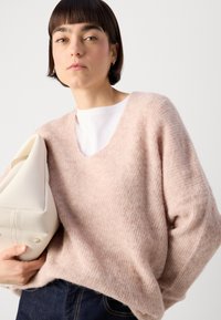 Helles Rosa, übergroßer Pullover aus weichem, strukturiertem Stoff mit V-Ausschnitt. Das Model hält eine cremefarbene Handtasche. Dunkelblaue Jeans sind sichtbar.
