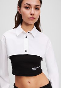 Cropped, two-tone shirt with a white top featuring a collar and black buttons, and a black ribbed bottom with "KARL LAGERFELD JEANS" embroidery.