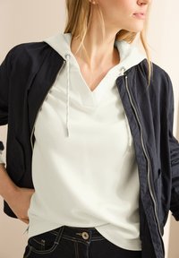 Marine bomberjack over een lichtgroene hoodie met een V-hals. Zacht materiaal, zilveren rits en details van de voorzak. Donkerblauwe jeans met gouden stiksel.