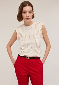Top bianco lavorato a maglia con maniche a volant, caratterizzato da un motivo a onde e dettagli a traforato, abbinato a pantaloni rossi e una cintura nera.