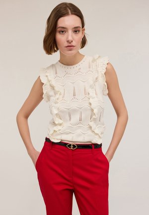 Weißes Stricktop mit Rüschensleeves, das ein Wellenmuster und durchbrochene Details aufweist, kombiniert mit roten Hosen und einem schwarzen Gürtel.