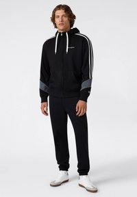 Felpa nera con zip e lacci bianchi, accentuata in grigio sulle maniche, indossata con pantaloni della tuta neri coordinati e sneakers bianche.