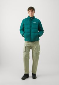 Teal pufjakke med quiltet design, høj krave og lynlås, parret med lysegrønne cargo-bukser og sorte sko.