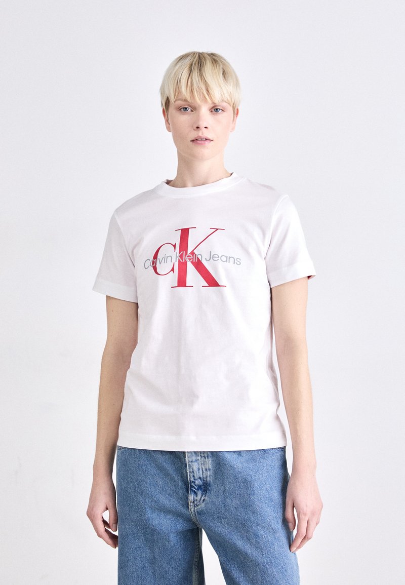Camiseta blanca de algodón con cuello redondo, que presenta un gran logo rojo "CK" y texto gris que dice "Calvin Klein Jeans". Textura suave.
