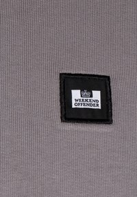 Tessuto grigio a coste con una toppa quadrata nera recante la scritta "WEEKEND OFFENDER" in bianco e un logo a forma di corona in alto.