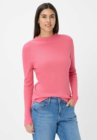 Geribbelde roze trui met lange mouwen en een hoge halslijn, gecombineerd met blauwe jeans. De textuur is zacht en de pasvorm is snug bij de taille.