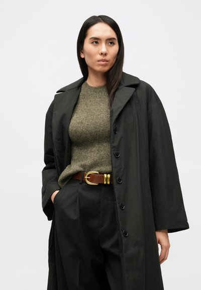 Čierny kabát po kolená s výrazným golierom a gombíkmi, v páre s olivovo zeleným pleteným svetrom a čiernymi nohavicami, doplnený hnedým opaskom so zlatou prackou.