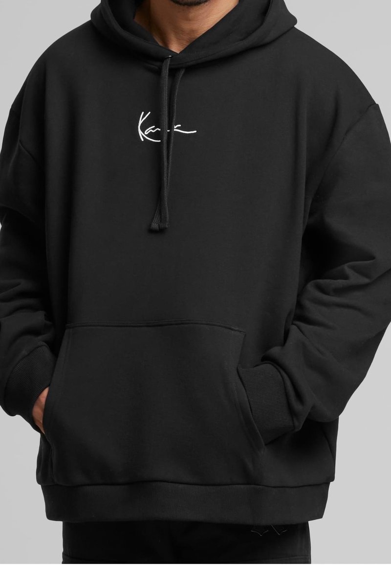 Homme portant un sweat à capuche noir avec un texte blanc de style signature sur la poitrine, mains dans la poche kangourou avant, sur un fond gris uni.