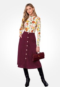 Mujer con blusa de manga larga floral, falda midi burdeos abotonada, botas negras hasta la rodilla, sosteniendo un bolso tejido burdeos.