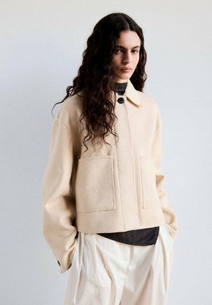 Mujer con cabello largo, oscuro y rizado, lleva una chaqueta beige clara sobre una camisa oscura y pantalones blancos de pierna ancha, de pie frente a un fondo liso.