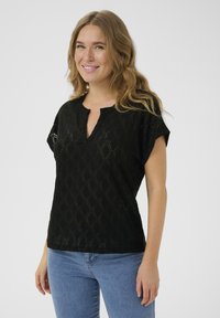 Black short-sleeve top with a v-neck and diamond pattern; made of lightweight material. Paired with blue denim jeans.