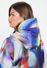 Chaqueta impermeable con capucha de colores, con un acabado liso y brillante, que presenta patrones abstractos en tonos de azul, púrpura y rojo.