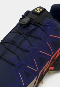 Sapatilha de corrida em azul-escuro com parte superior texturizada, detalhes em laranja contrastante e atacadores elásticos. Apresenta um ajuste seguro com um mecanismo de bloqueio.