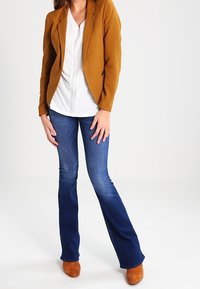 Blazer senape sopra una blusa bianca, abbinato a jeans blu a zampa d'elefante e stivaletti marroni. Tessuto liscio con vestibilità su misura e dettagli sottili.