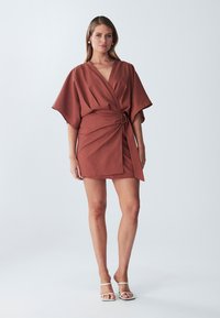 Haljina u boji hrđe s dizajnom omota, kratke duljine, kimono rukavima i vezanom akcentom u struku, izrađena od teksturirane tkanine.