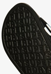 Suola nera di calzature con un motivo a rilievo del marchio "SUPERDRY", caratterizzata da una finitura liscia e una forma arrotondata, che mette in risalto la sua texture e il branding.
