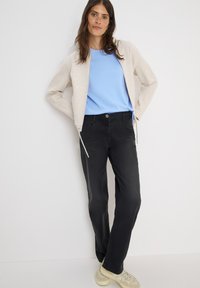 Lichtbeige zip-up jacket, blauwe t-shirt, zwarte broek en lichte sneakers. De jas heeft een aangesloten ontwerp; de t-shirt is met ronde hals; de broek heeft een rechte pijp.