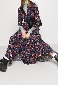 Lang, patroon jurk in donkerblauw met een roze en gele luipaardprint; gecombineerd met een groene coltrui en een zwarte schoudertas. Zwarte laarzen maken de look compleet.