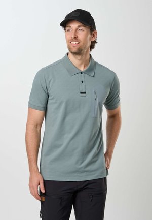Homme portant un polo vert clair avec une poche poitrine zippée et une casquette de baseball noire, regardant sur le côté et souriant légèrement.