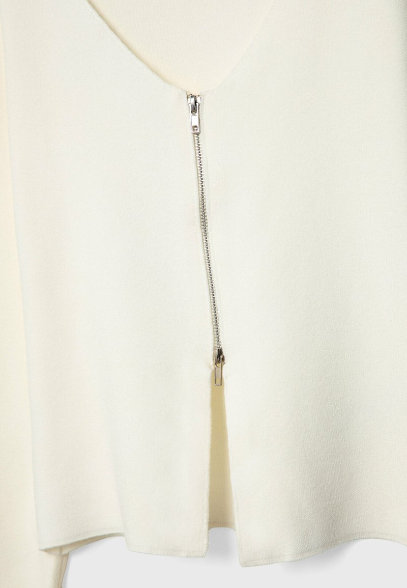 Indumento color crema con scollatura a V, dotato di una cerniera argento sul davanti. Tessuto morbido e orlo leggermente svasato.