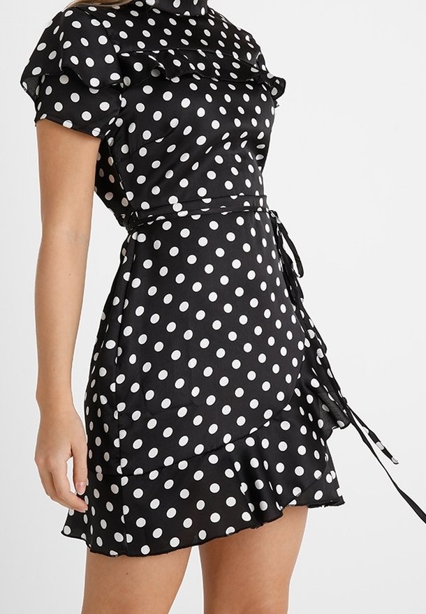 Robe portefeuille noire à manches courtes avec des pois blancs, ourlet à volants et ceinture à la taille, portée par une personne avec un bras visible.