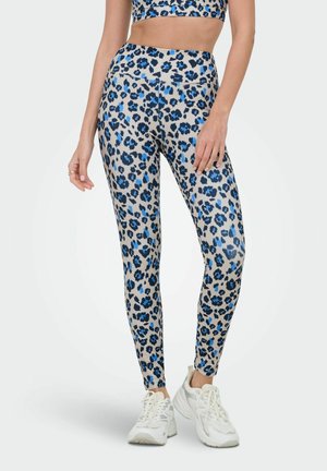Femme portant des leggings beige à taches léopard bleues et noires, associés à des baskets blanches, debout devant un fond uni.