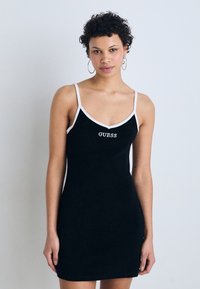 Vestido negro ajustado con tirantes finos, ribete blanco y logo "GUESS" en el pecho. La tela parece suave, con una textura lisa.