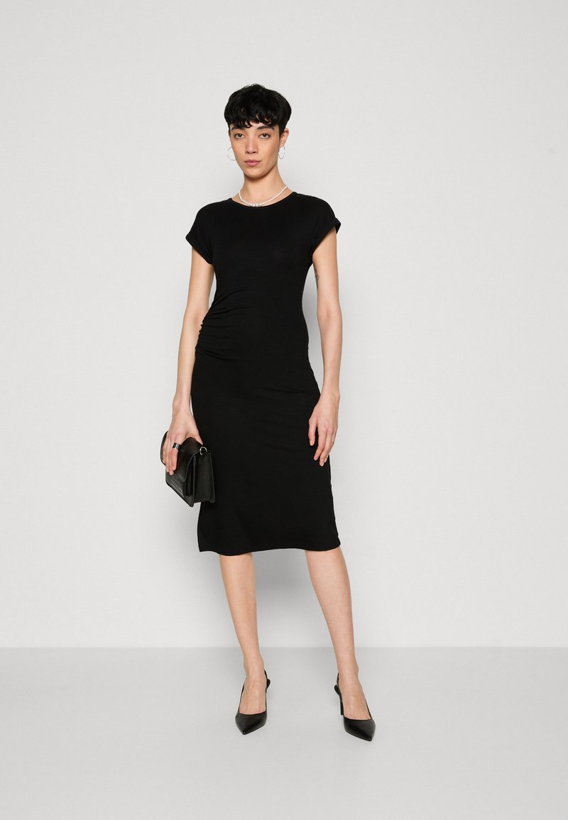 Czarna dopasowana sukienka z krótkimi rękawami i marszczeniem po bokach. Modelka trzyma małą czarną torebkę, ma na sobie czarne szpilki. Prosta, gładka tekstura.