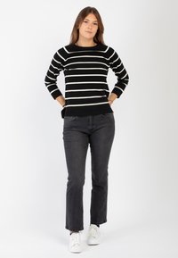 Maglione a righe bianche e nere con maniche lunghe e scollatura rotonda, abbinato a jeans scuri e sneakers bianche, ambientato su uno sfondo neutro.