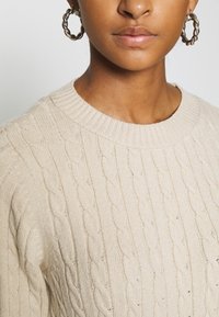 Maglione beige a lavorazione cable con collo a costine. Presenta una texture morbida con dettagli sottili. La modella indossa orecchini a cerchio in argento.