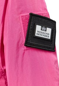 Lyserød letvægtsjakke med en tekstureret lap med sort kant og en rektangulær label, der står "WEEKEND OFFENDER." Glat stofstruktur.
