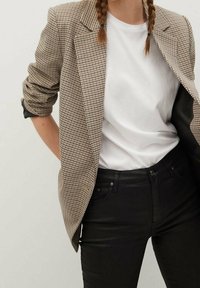 Blazer de espiga marrón y negro con solapas recortadas, llevado sobre una camiseta blanca, combinado con jeans ajustados negros. Textura suave.
