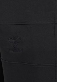 Pantaloni sportivi neri con finitura testurizzata, una linea di cucitura visibile e un logo in rilievo sulla parte anteriore inferiore.