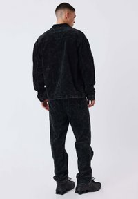 Uomo in piedi di spalle, indossando una giacca di denim nera lavata e pantaloni con sneakers nere, su uno sfondo grigio chiaro uniforme.