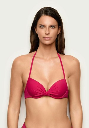 Femme aux longs cheveux bruns portant un haut de bikini rose vif dos-nu, face à un arrière-plan clair et uni.