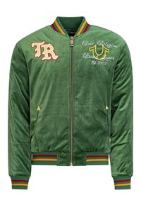 Groene fluwelen bomberjack met geribbelde kraag en boorden. Beschikt over crème en gele borduuring, ritssluiting en twee zijzakken.